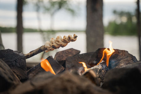 Pinnbröd grillas över öppen eld med en sjö och skog i bakgrunden.
