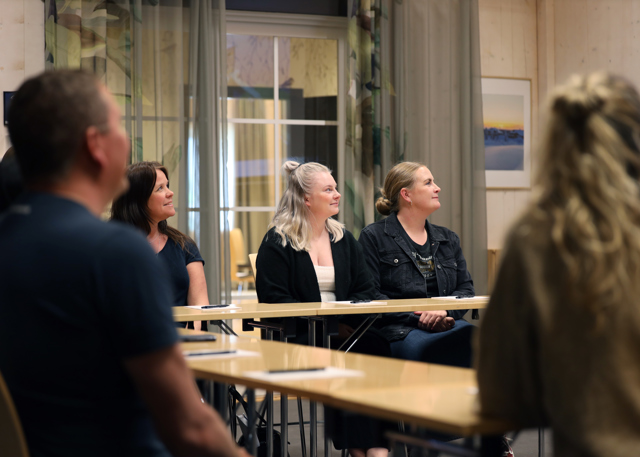 Vuxna sitter vid bord i ett mötesrum och lyssnar under en presentation.