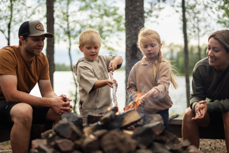 En familj sitter runt en lägereld vid en sjö, där två barn grillar pinnbröd medan de vuxna tittar på och ler.