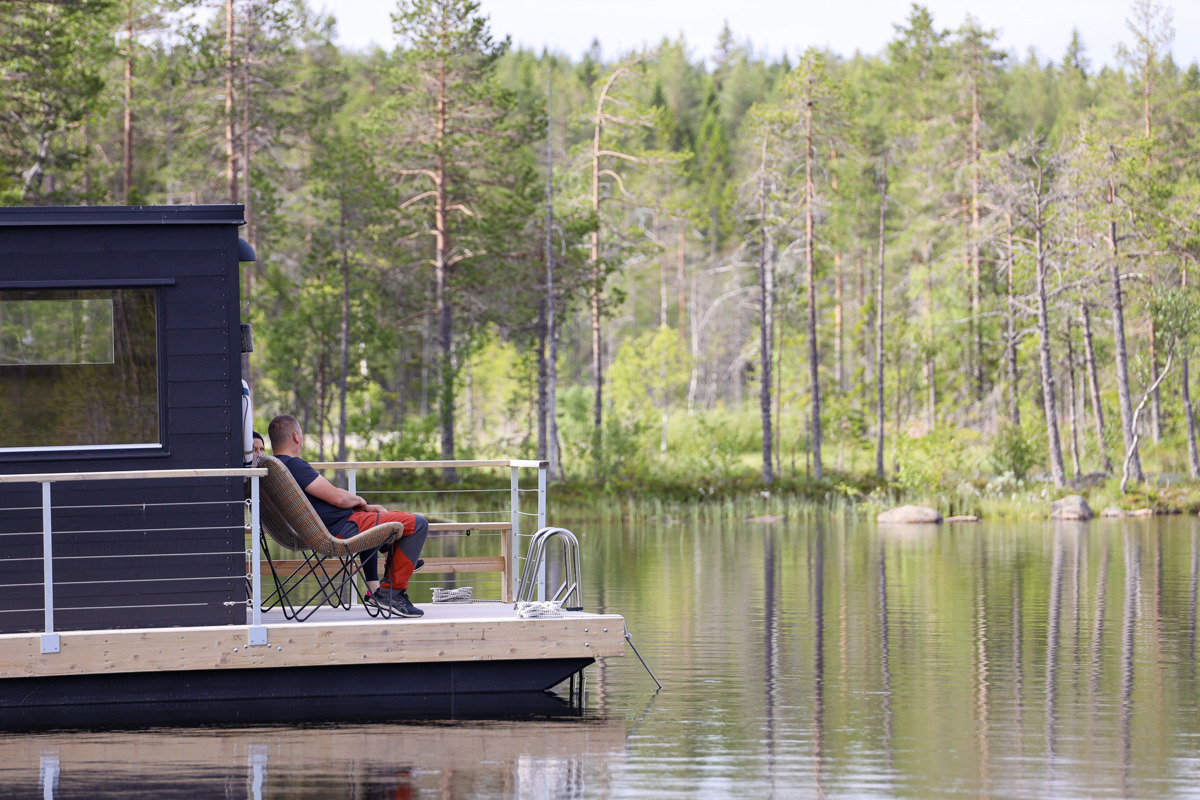 Person sitter i stol på en bastuflotte vid en skogssjö.