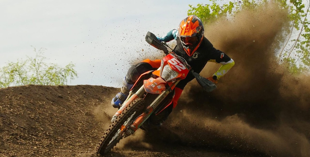 Motocrossförare i orange hjälm kör i hög fart genom en kurva, med jord som yr upp bakom motorcykeln.