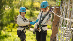 Ett barn och en vuxen med hjälm och sele ler tillsammans i klätterparken, fastkopplade i säkerhetslinan.