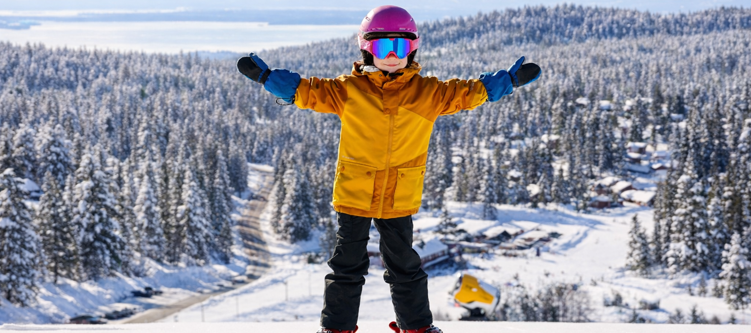 Ett barn står på skidor med armarna utsträckta i en snöklädd skidbacke, med snötäckta skogar och bebyggelse i bakgrunden.